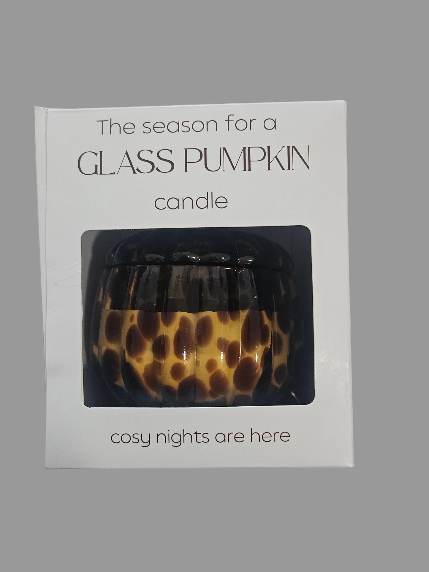 A boxed glass pumpkin candle shown on a plain light background. The white package has printed text reading 'The season for a GLASS PUMPKIN candle' and 'cosy nights are here,' with a window revealing the candle inside.