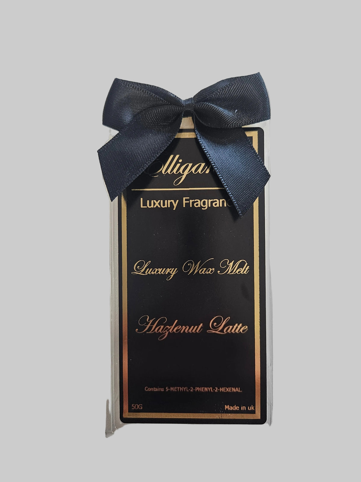 A packaged luxury fragrance wax melt in a clear plastic clamshell with a dark navy ribbon bow. The insert card is black with gold lettering reading 'Alligan Luxury Fragran', 'Luxury Wax Melt', and 'Hazelnut Latte'.