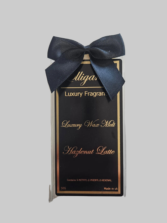 A packaged luxury fragrance wax melt in a clear plastic clamshell with a dark navy ribbon bow. The insert card is black with gold lettering reading 'Alligan Luxury Fragran', 'Luxury Wax Melt', and 'Hazelnut Latte'.